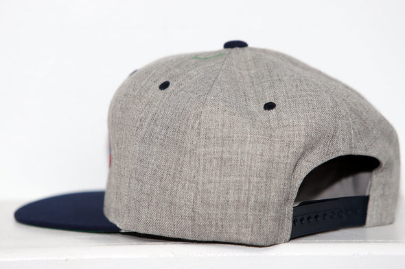 Side view of a gray and navy Wichita Wind snapback hat with adjustable strap on white surface